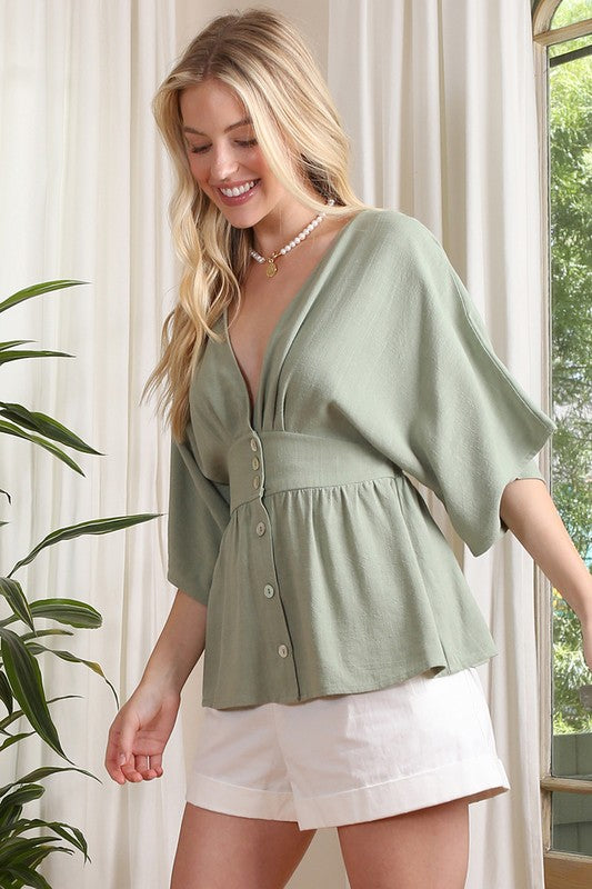 A woman in an SS deep V neck button-down top and a white skirt stands near a potted plant in a room with large windows.