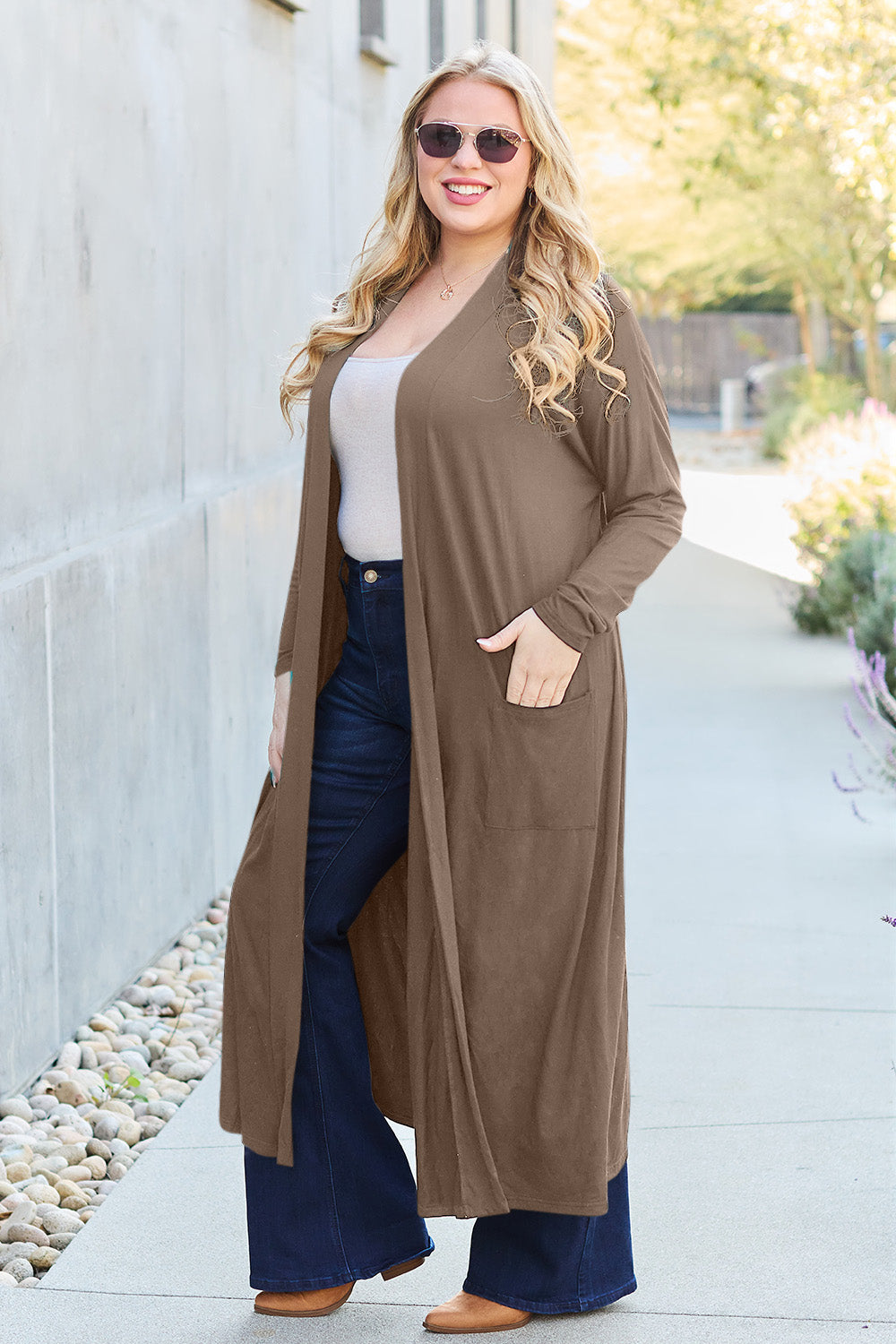 A woman stands against a concrete wall, wearing the Basic Bae Full Size Open Front Long Sleeve Cover Up made from rayon spandex, a white top, blue jeans in her perfect size, and brown shoes with her hands in her pockets. Machine wash cold for best results.