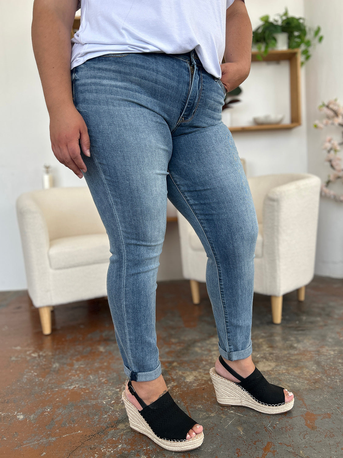 A person wearing a white shirt, Judy Blue Full Size Cuffed Hem Slim Jeans, and black wedge-heeled sandals is standing in a room with beige chairs and plants in the background.