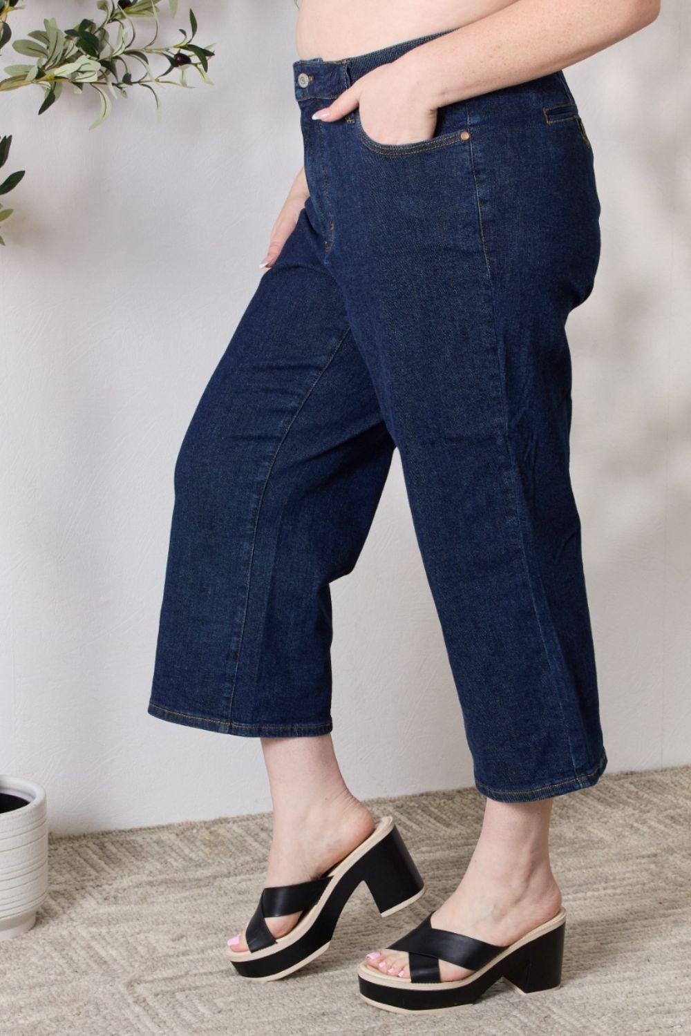 A person wearing the Judy Blue Full Size High Waist Cropped Wide Leg Jeans, which are renowned for their tummy control design, and nude block heel sandals stands near a potted plant.