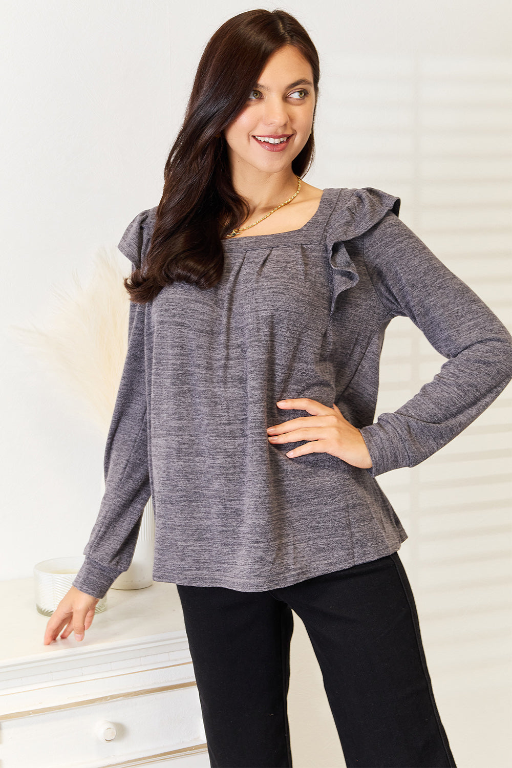 A woman wearing a Double Take Square Neck Ruffle Shoulder Long Sleeve T-Shirt in pink and blue jeans stands with one hand in her pocket, smiling. She has long curly hair and wears a necklace and bracelet. She stands in a room with white decor.