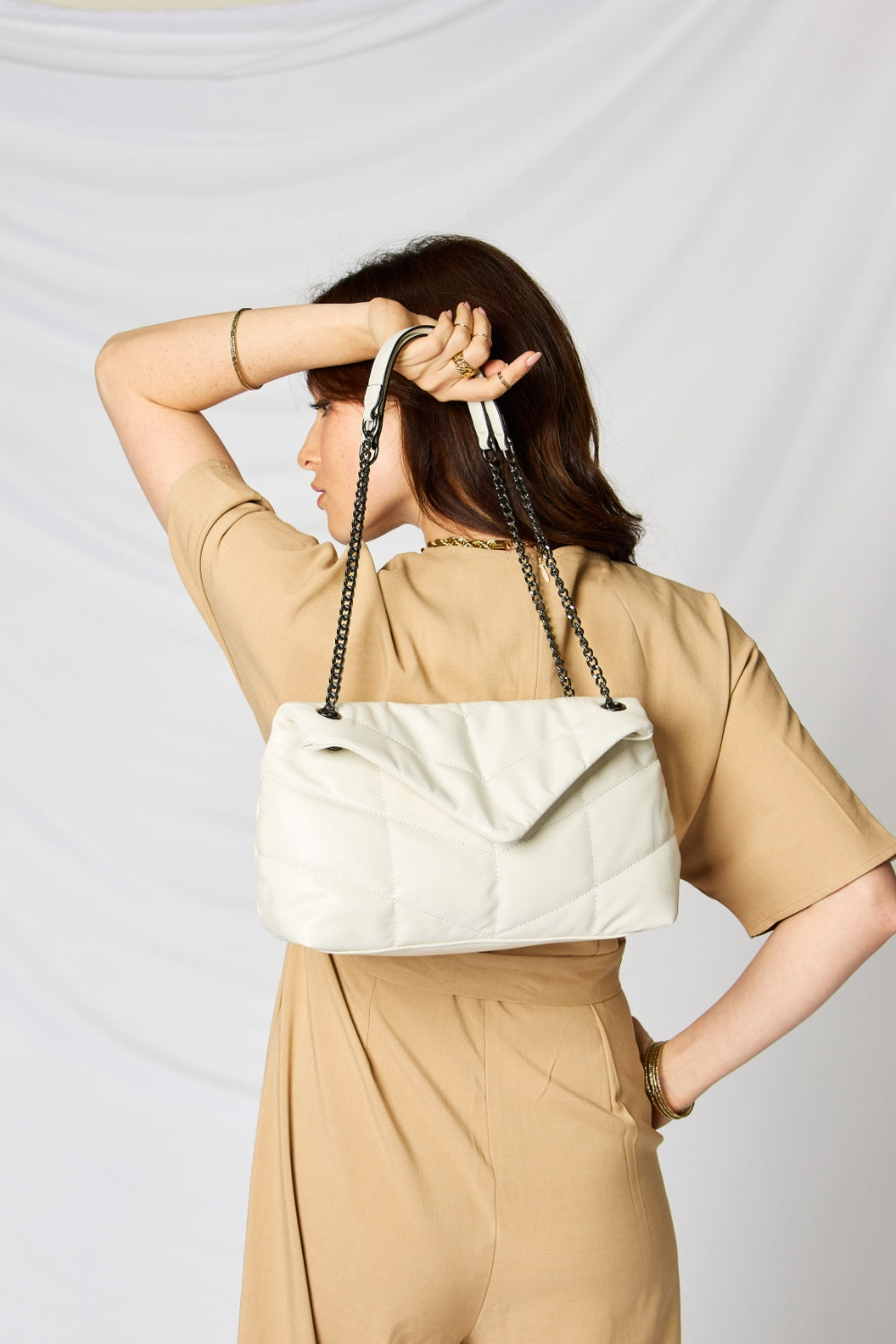 A person holding a stylish SHOMICO PU Leather Chain Handbag over their shoulder against a white background. They are dressed in a beige outfit and looking at the camera, embodying sustainable fashion.