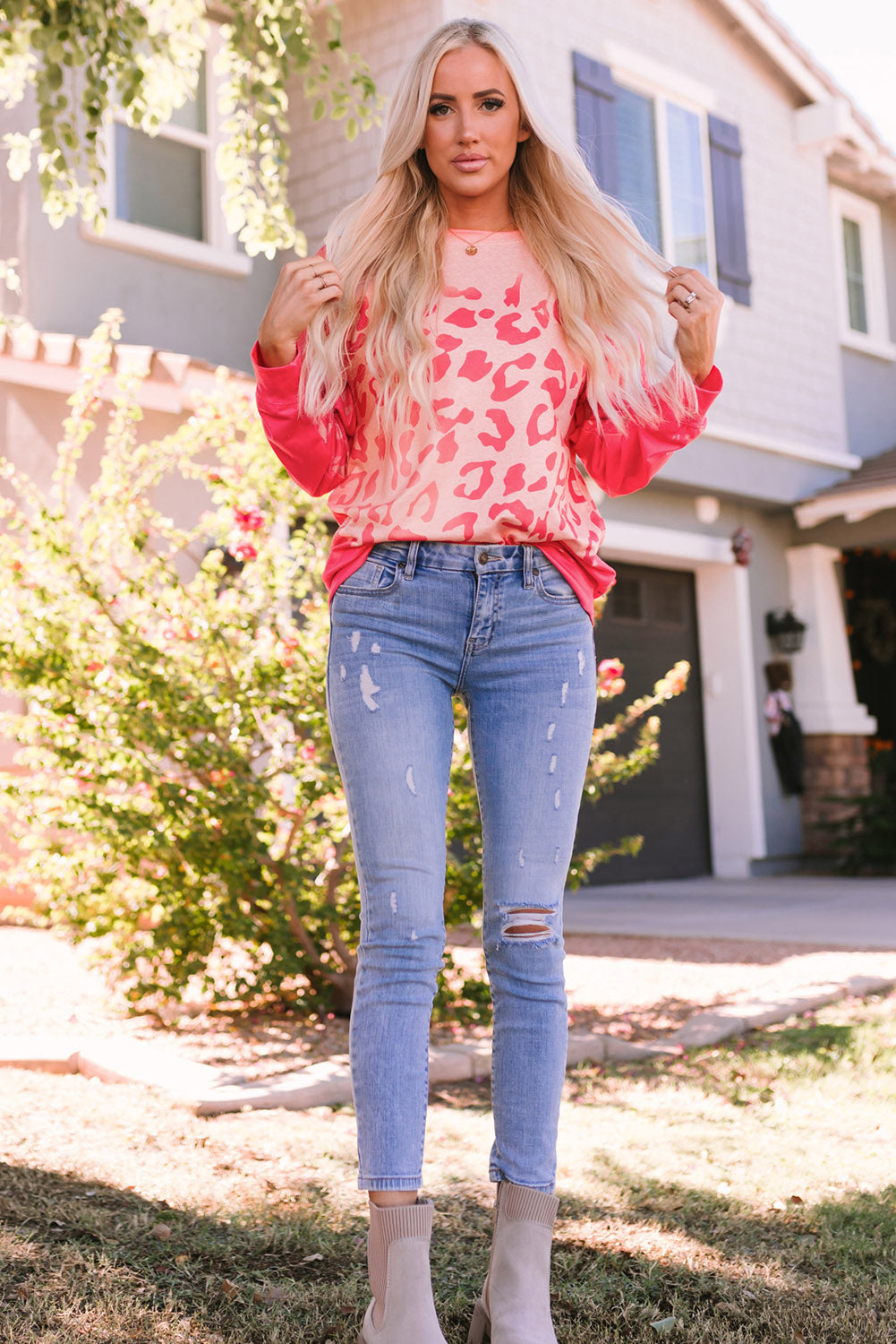 A woman with long blonde hair stands outdoors wearing the Pink Bleached Cheetah Print Sweatshirt and blue jeans. She is smiling and posing with one hand in her pocket.