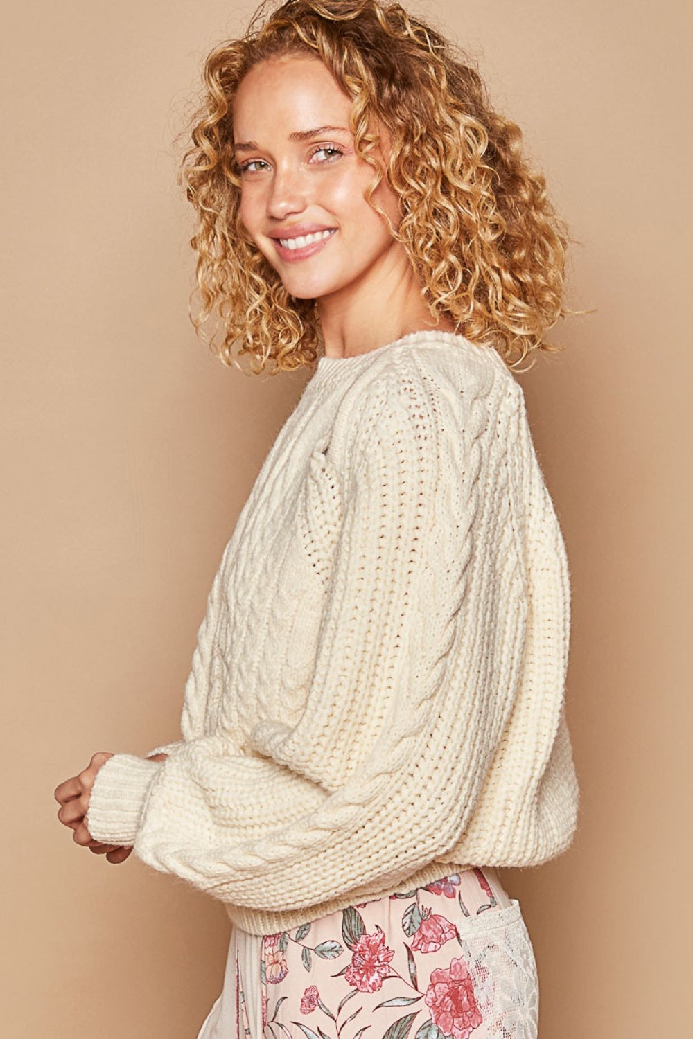 A person wearing the POL Cable Knit Cutout Long Sleeve Sweater in cream, paired with a stylish plaid skirt, stands outdoors surrounded by lush green foliage.