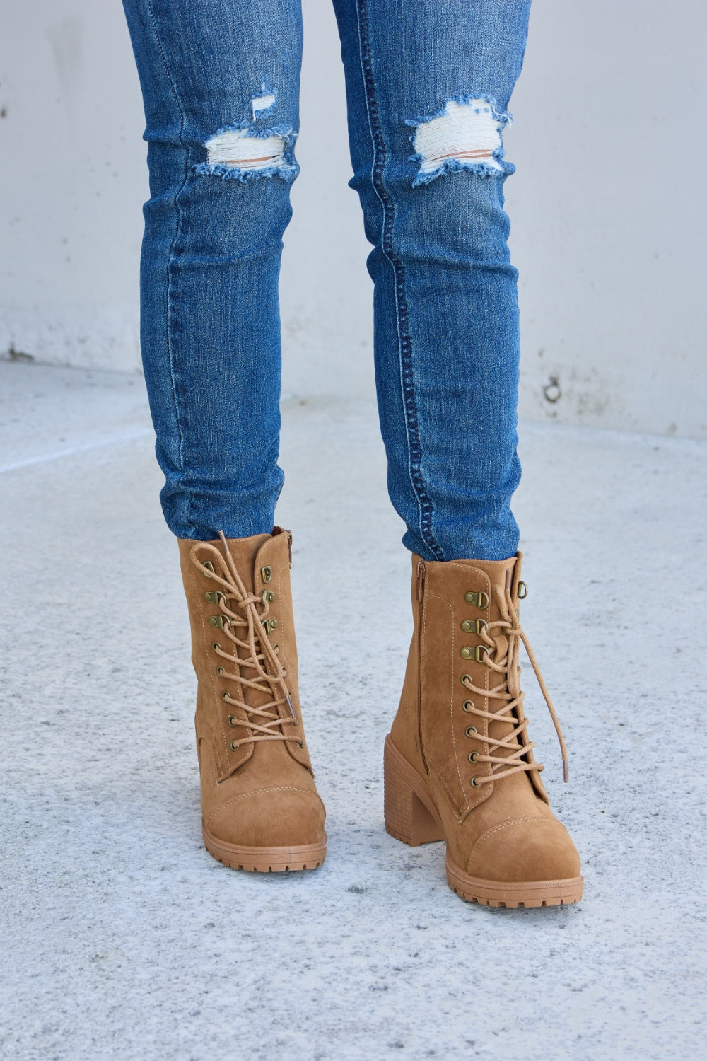 A person wearing distressed blue jeans and Forever Link Lace-Up Zipper Detail Block Heel Boots stands on a concrete surface.