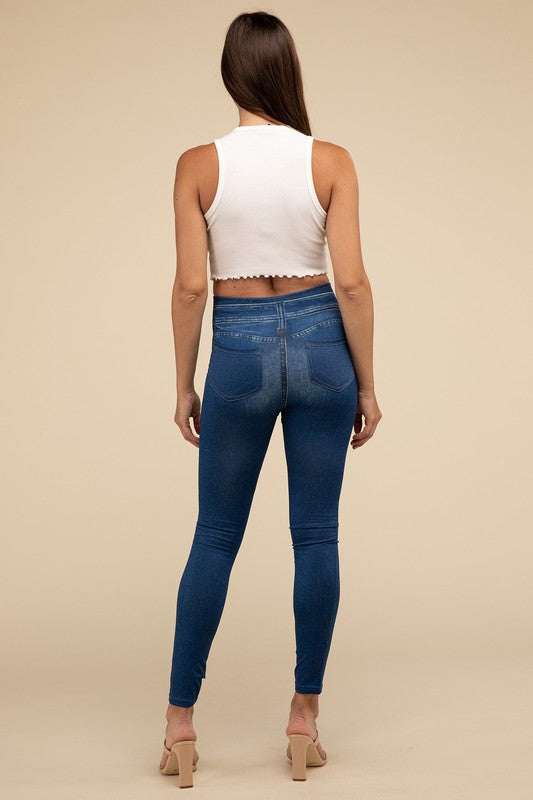 A person wearing Denim Leggings in a high-waisted black design and paired with black heels poses against a plain beige background, highlighting the sleek, stretchy fit.