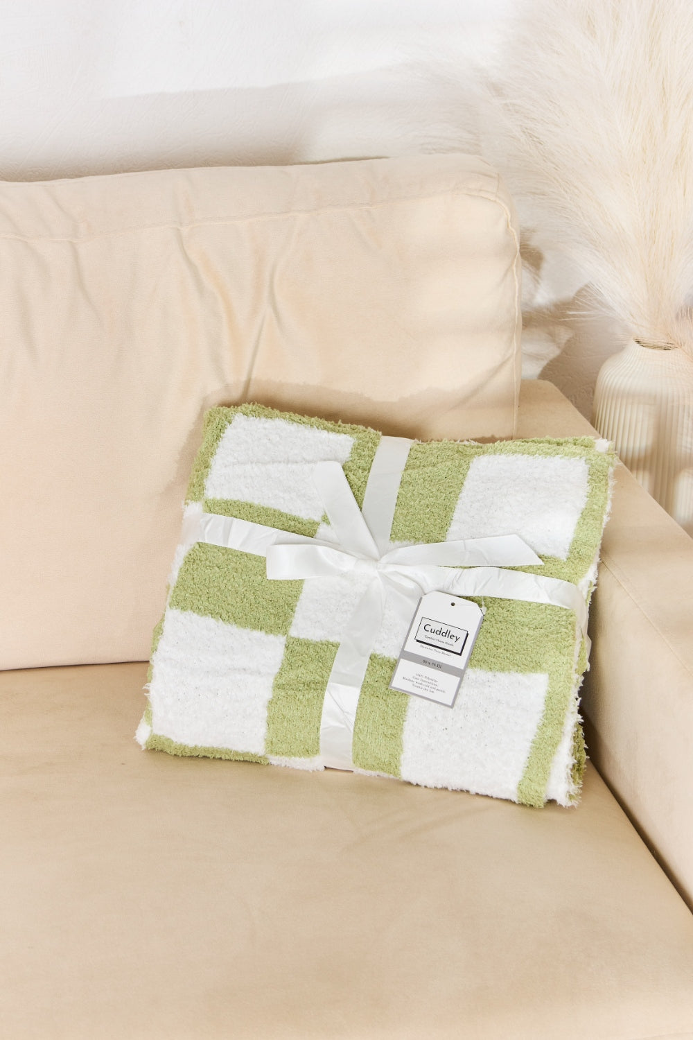 A person stands holding a large Cuddley Checkered Decorative Throw Blanket, smiling, in a room with a sofa and a potted plant. The blanket features a black and white checkerboard pattern, is made from 100% polyester, and imported for quality.