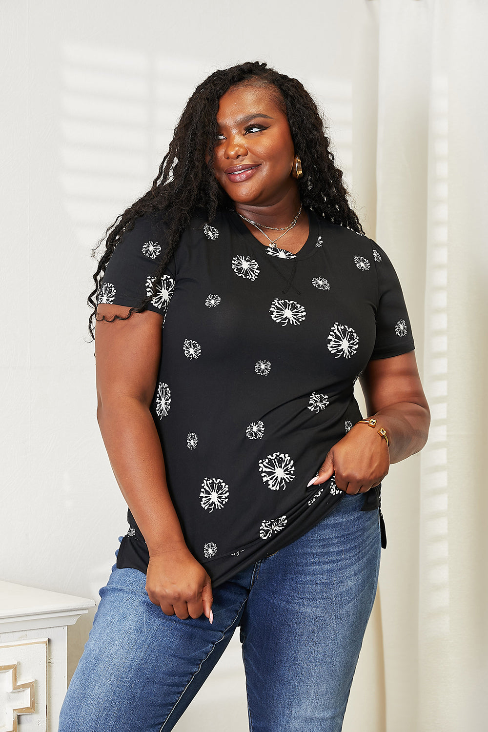 A woman with long braided hair is smiling and wearing the Double Take Dandelion Print Round Neck T-Shirt paired with blue jeans. She stands in front of a light-colored wall, embodying nature-inspired fashion.