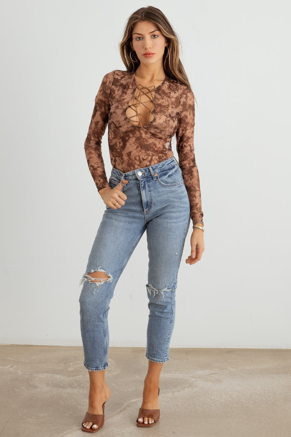 A woman with long hair is wearing the HERA COLLECTION Abstract Mesh Lace-Up Long Sleeve Bodysuit paired with ripped blue jeans, creating a statement-making look as she stands against a plain background.