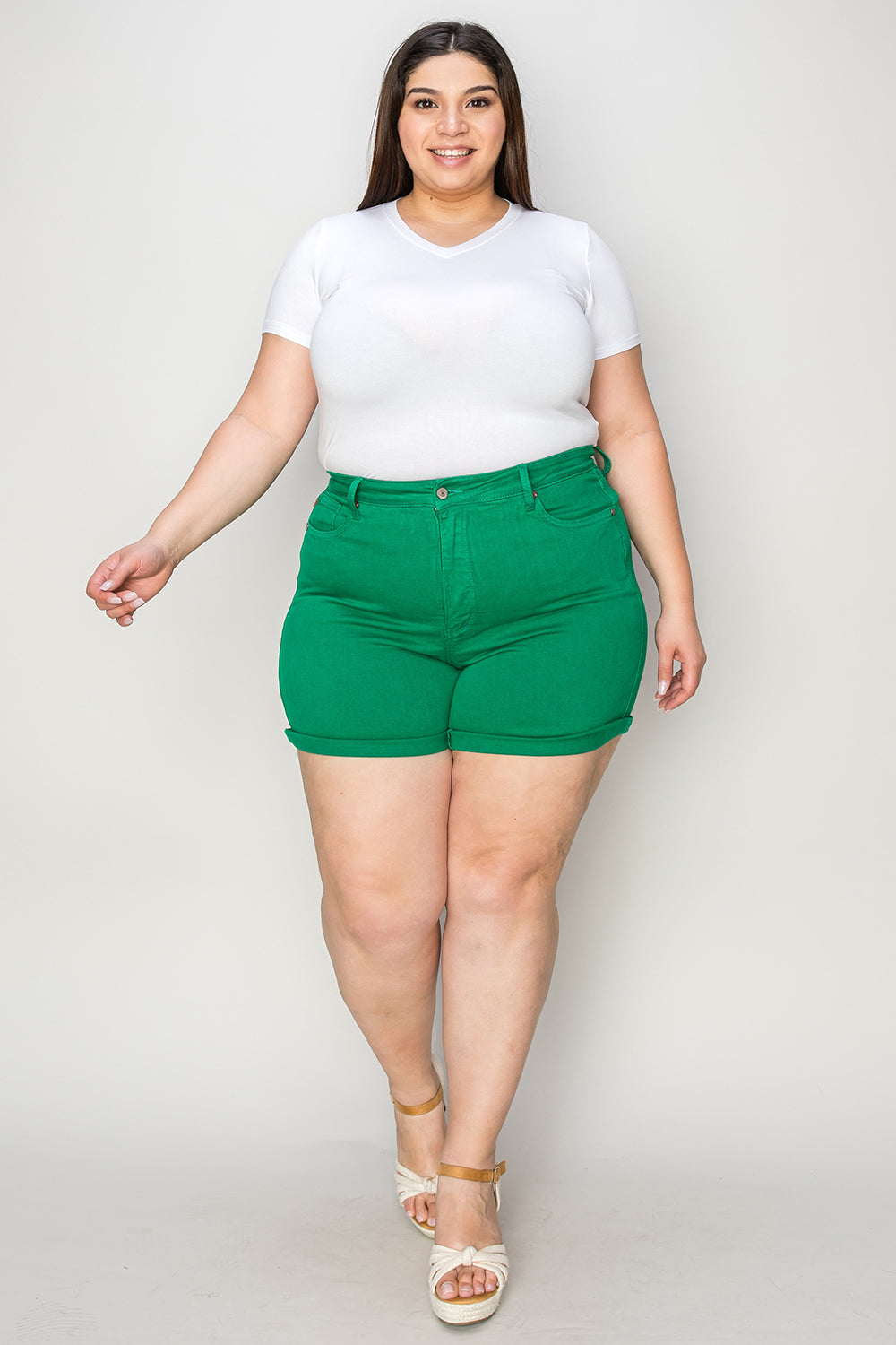 Two women stand indoors wearing white tops and Judy Blue Full Size Tummy Control Garment Dyed Denim Shorts in green. The woman on the left has her hand in her pocket, and both are smiling. There are plants and decorations in the background.