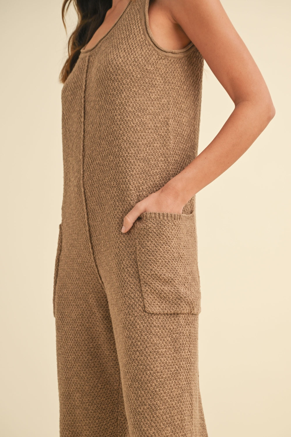 A woman stands against a beige background wearing the MABLE Sleeveless Knit Crop Jumpsuit with Pockets in brown and beige shoes.