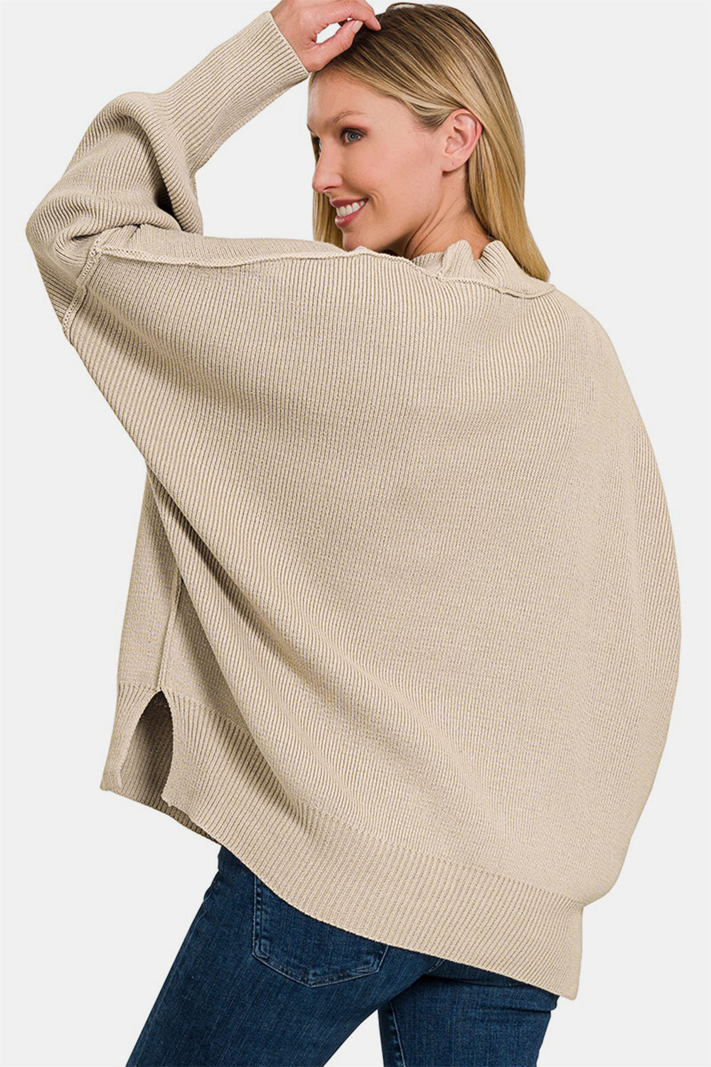 A person wearing the Zenana Side Slit Oversize Sweater, a cozy knit in beige, paired with jeans, smiling with one hand touching their hair.