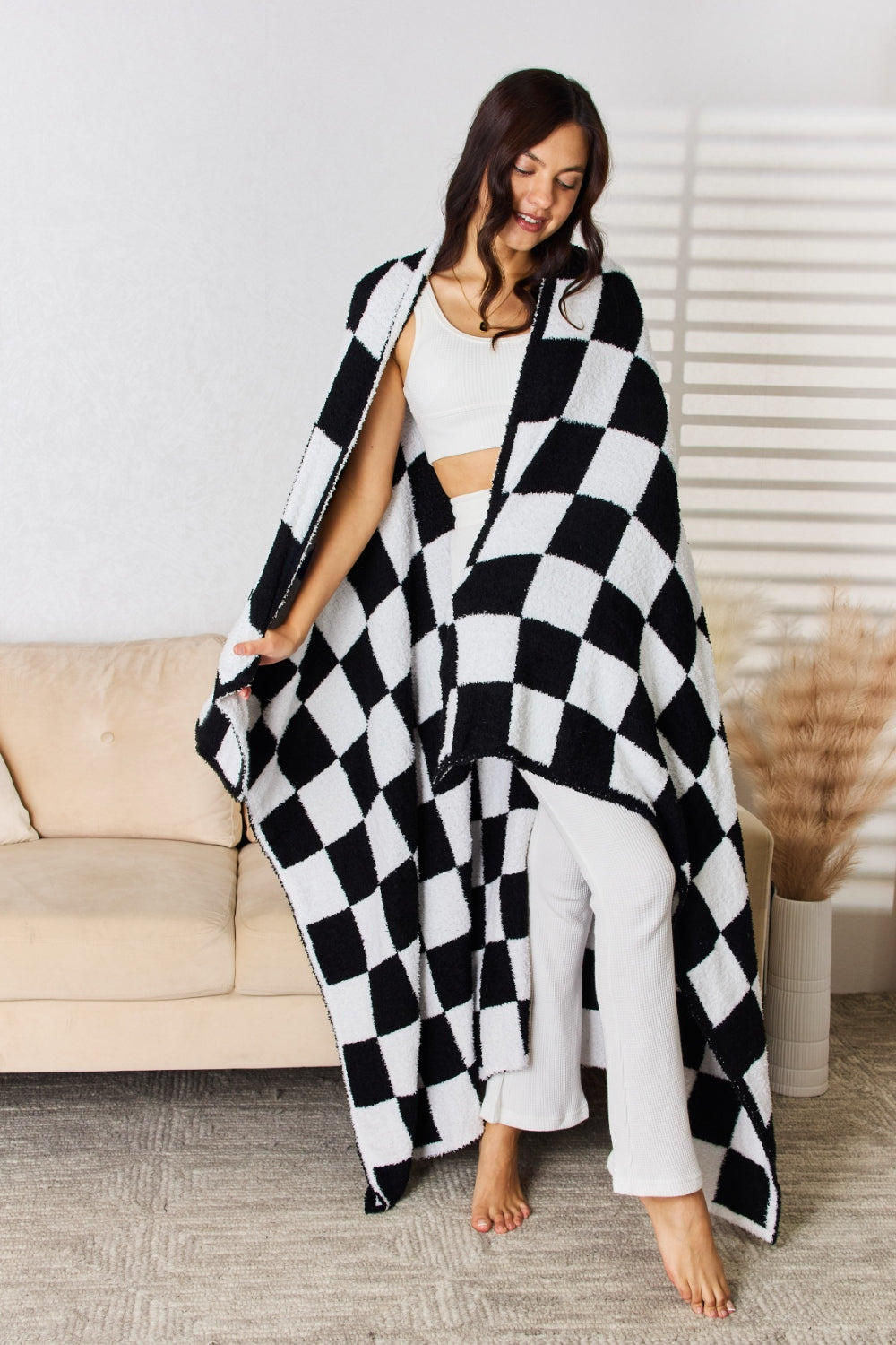 A person stands holding a large Cuddley Checkered Decorative Throw Blanket, smiling, in a room with a sofa and a potted plant. The blanket features a black and white checkerboard pattern, is made from 100% polyester, and imported for quality.