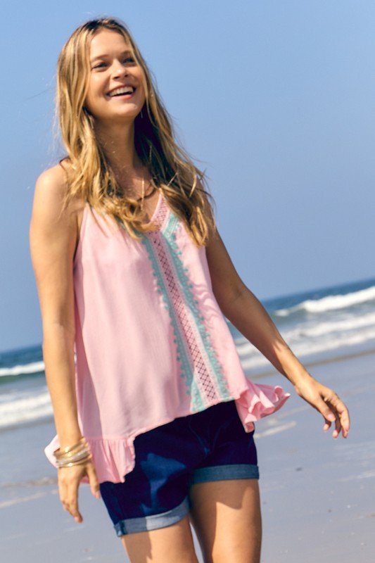 A woman with long hair is wearing a Printed Sleeveless Ruffle Tank Top in light blue and pink, paired with denim shorts, as she stands against a neutral background.