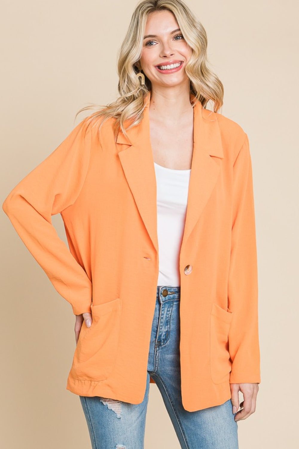 A smiling person models the Culture Code One Button Long Sleeve Blazer with Pockets, a tailored fit in vibrant orange, over a white top and blue jeans. They pose with one hand in their pocket against a plain background, perfectly capturing versatile fashion in this stylish look.