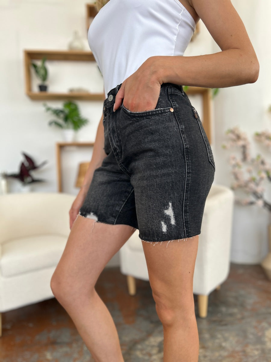 A person wearing a white t-shirt and Judy Blue Full Size High Waist Rigid Magic Denim Shorts is standing indoors in front of a beige chair.