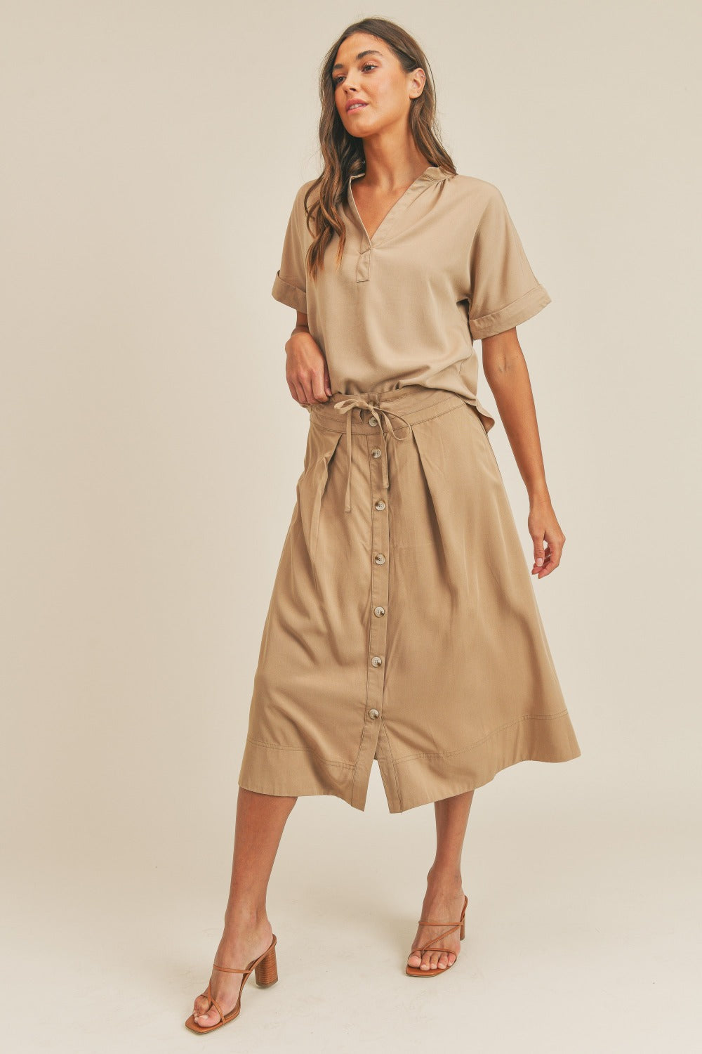 A woman is standing against a plain background wearing the MABLE Short Sleeve Top and Button Down Midi Skirt Set in beige. The elegant two-piece set, which includes a belted midi skirt, is paired perfectly with beige heels as she confidently poses with one hand on her hip.