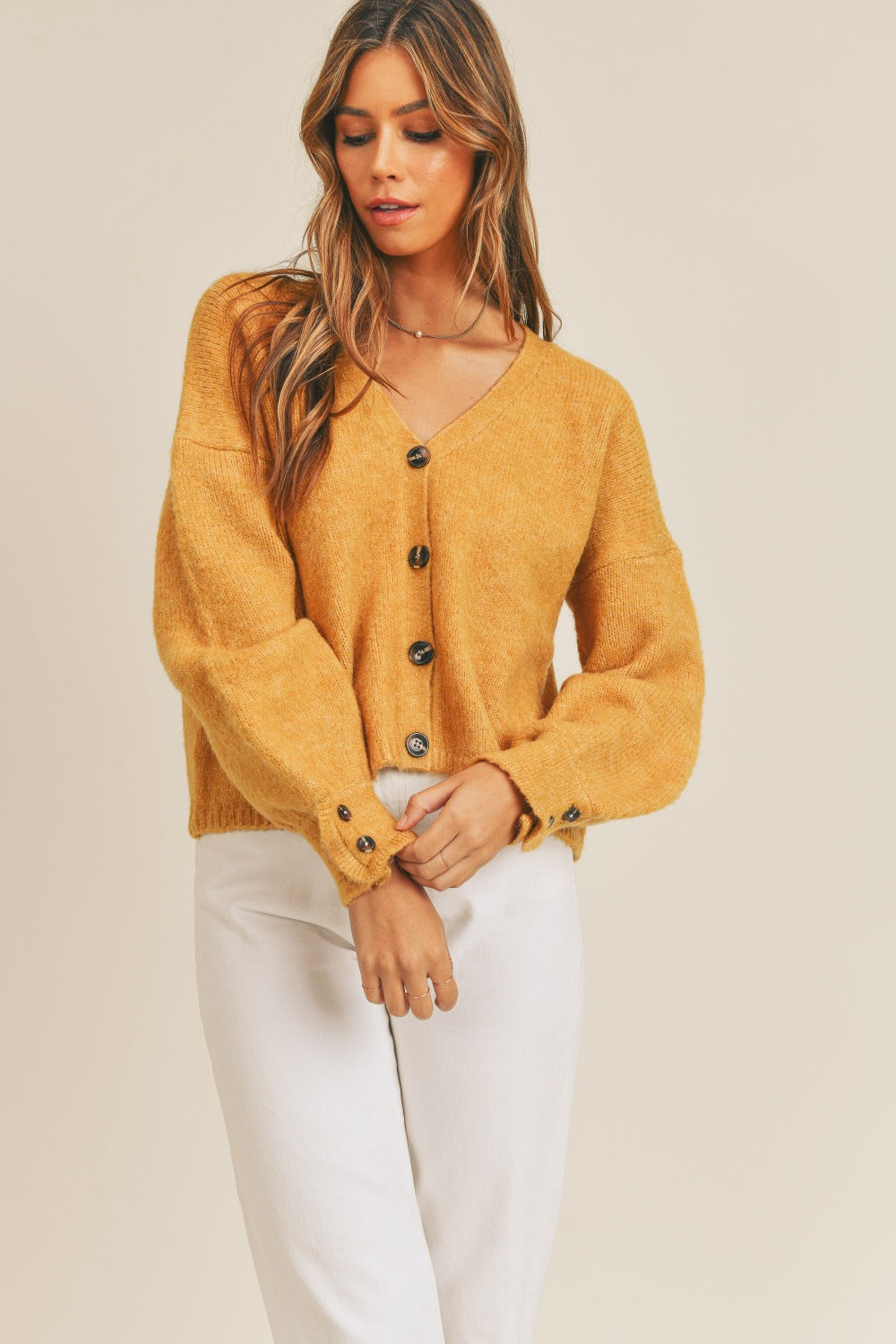 A person wears the MABLE Long Sleeve Button Down Sweater Cardigan in mustard, paired with white high-waisted pants, standing in front of a plain background.