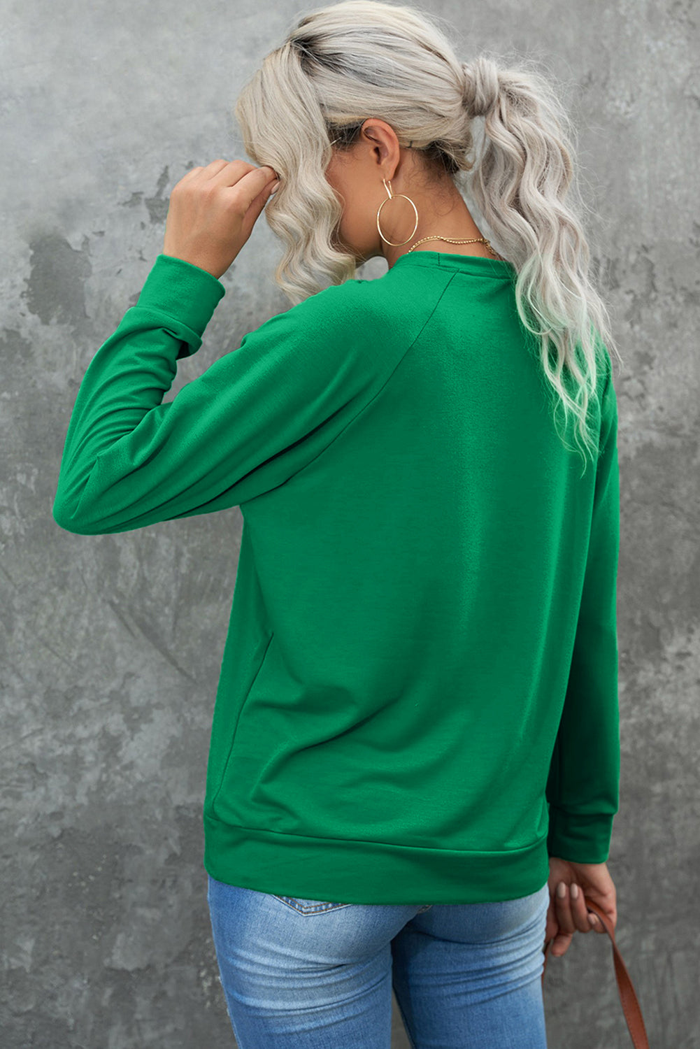 A person with long, light hair is standing with their back to the camera, wearing a comfy Green Solid Round Neck Raglan Sleeve Sweatshirt and blue jeans, against a gray textured wall background.