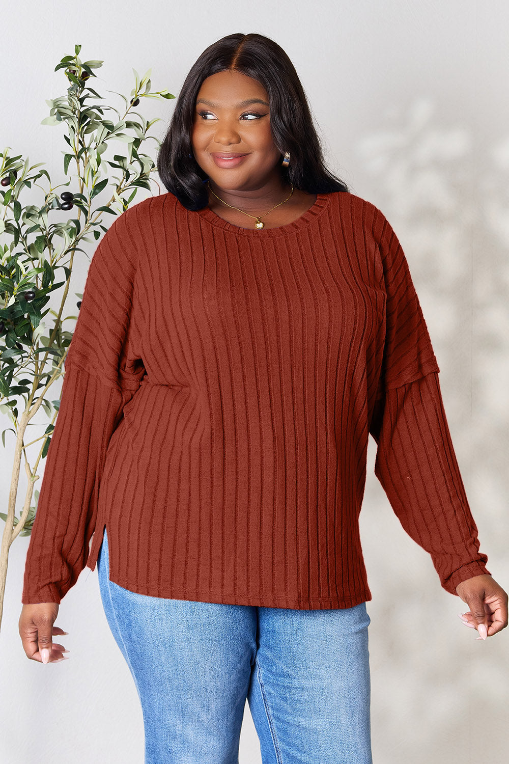 A person wearing the Basic Bae Full Size Ribbed Round Neck Slit Knit Top in blue and beige pants stands and smiles in front of a leafy plant.