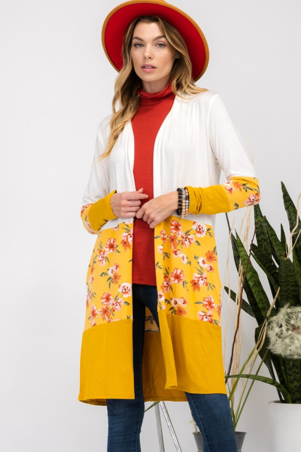 Wearing the Celeste Full Size Floral Color Block Open Front Cardigan with a white and yellow floral print, along with a red hat, a red top, and jeans, a woman stands beside a houseplant.