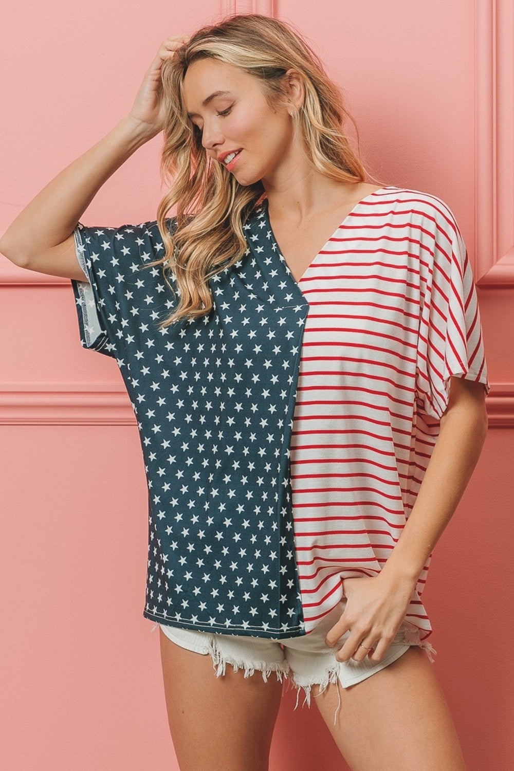 A woman stands against a pink wall, wearing the BiBi US Flag Themed Color Block Short Sleeve T-Shirt, which features one half blue with white stars and the other half red and white stripes. She is smiling with long wavy hair, showcasing her American pride casual wear.