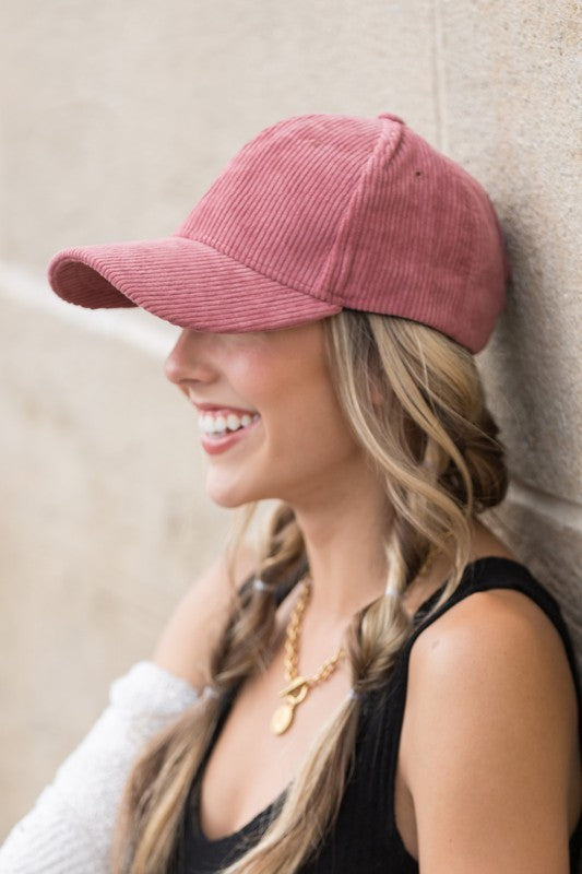 Sentence with the replaced product name: Woman smiling and leaning against a wall while holding the brim of her Corduroy Boyfriend Ball Cap.