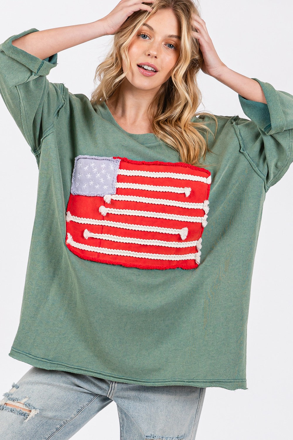 A person exudes casual charm in the SAGE + FIG Full Size American Flag Patch Drop Shoulder T-Shirt, paired with ripped jeans, confidently posing with hands in hair against a plain background.