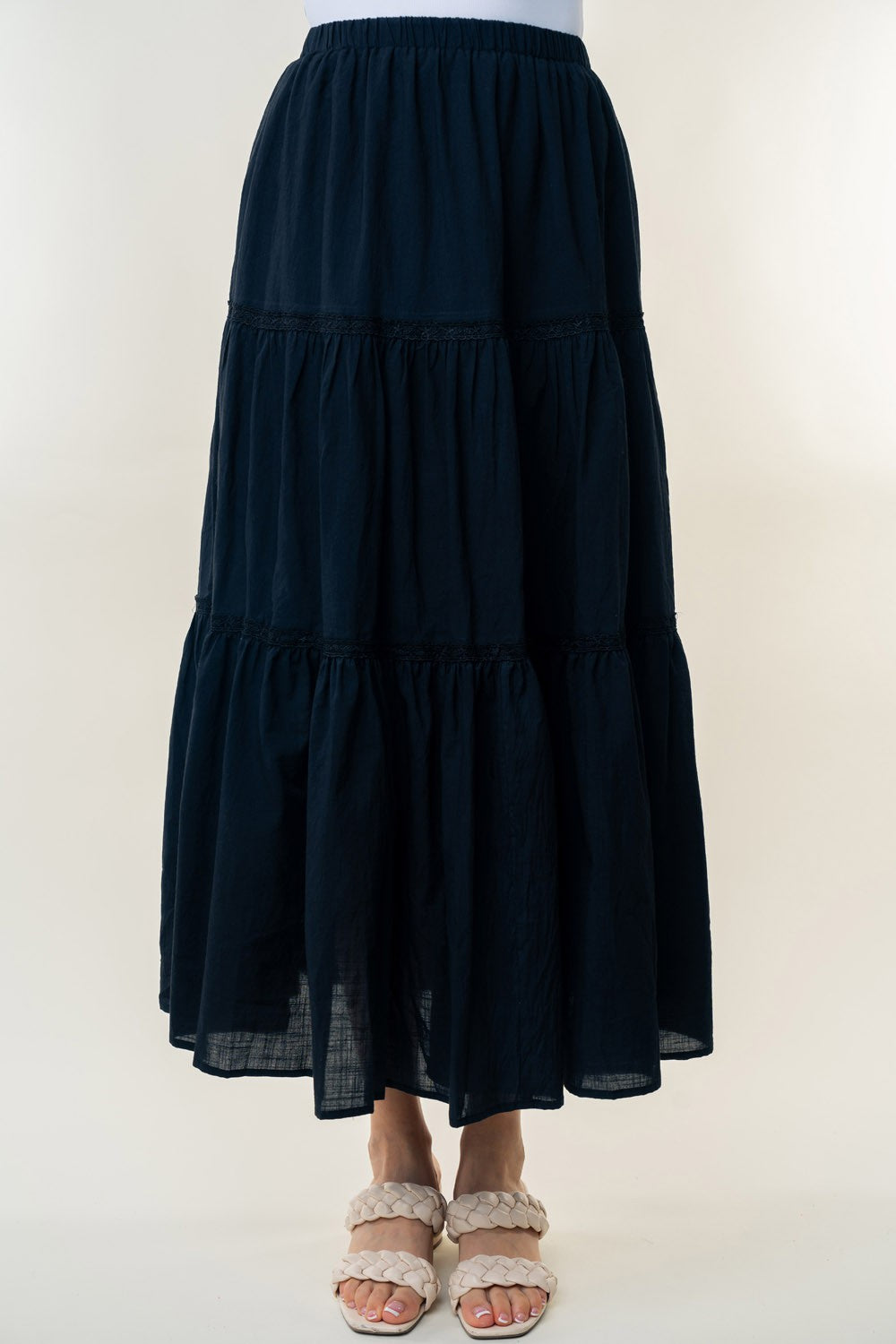 A long, White Birch High Waisted Tiered Maxi Skirt in navy blue, offering a versatile piece perfect for a bohemian look. It's worn by a person paired with white heeled sandals. The background is plain and light-colored.