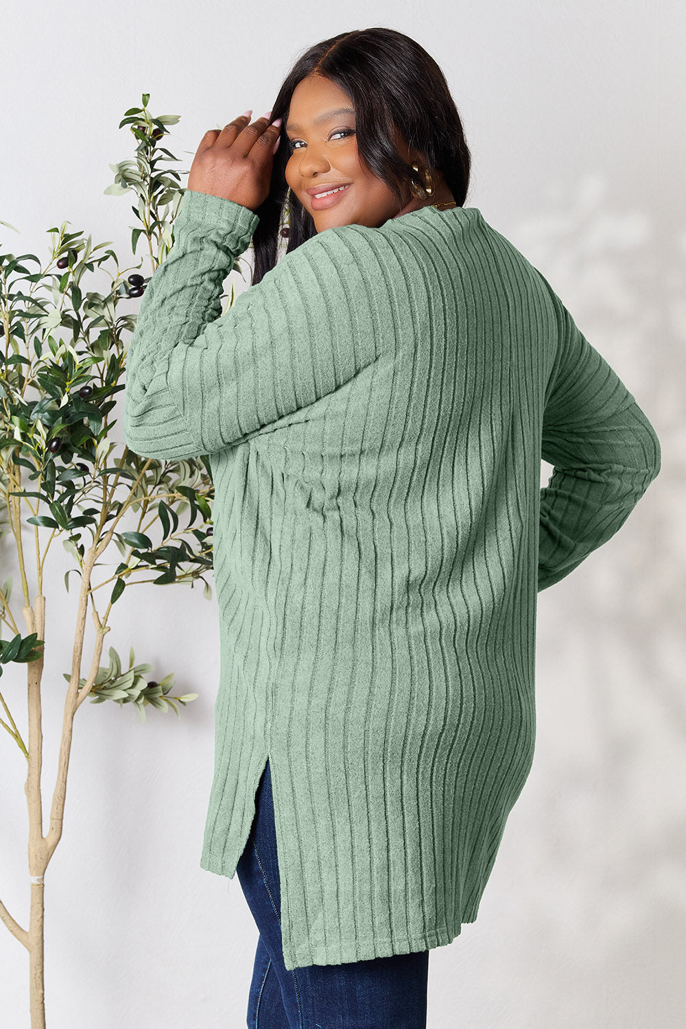 A person with shoulder-length hair smiles while wearing a beige ribbed sweater called the Basic Bae Full Size Ribbed Round Neck Long Sleeve Slit Top, along with matching beige pants, showcasing a basic style. They stand next to a green plant.