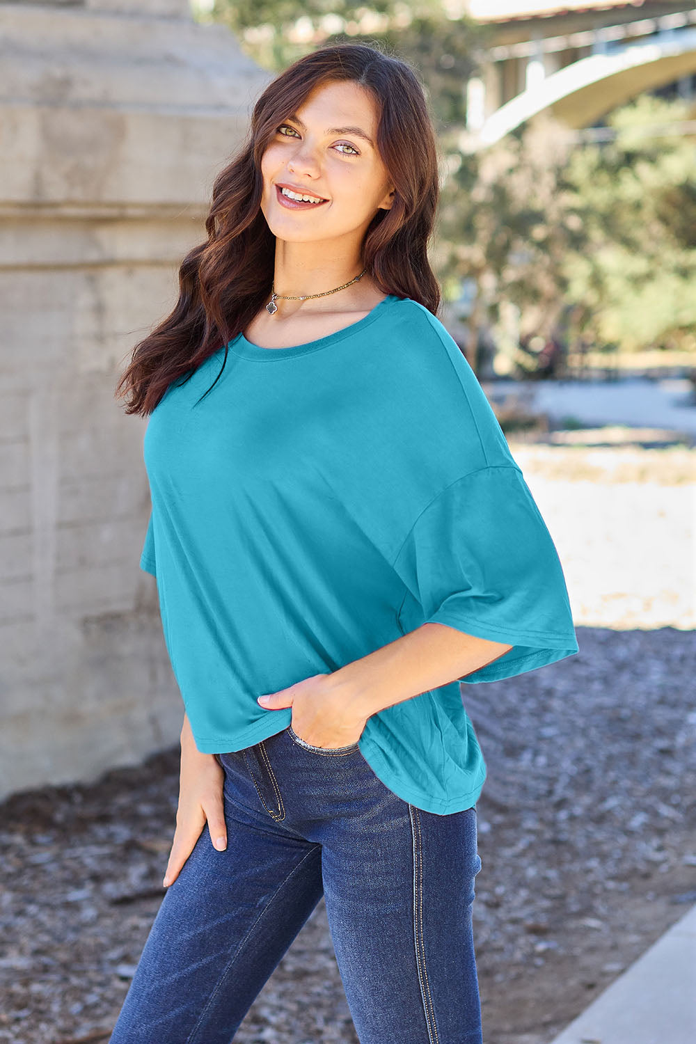 A person with long dark hair and a bright smile stands outdoors, showcasing their basic style in a loose, slightly stretchy *Basic Bae Full Size Round Neck Drop Shoulder T-Shirt* in pink and blue jeans, with one hand on their hip.