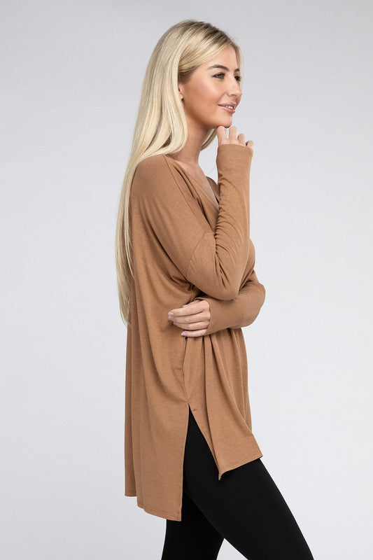 A woman with long, blonde hair is wearing a loose-fitting, brown Dolman Long Sleeve V-Neck Side Slit Hi-Low Hem Top and black pants. She poses with one hand on her hip and a neutral facial expression against a light grey background.