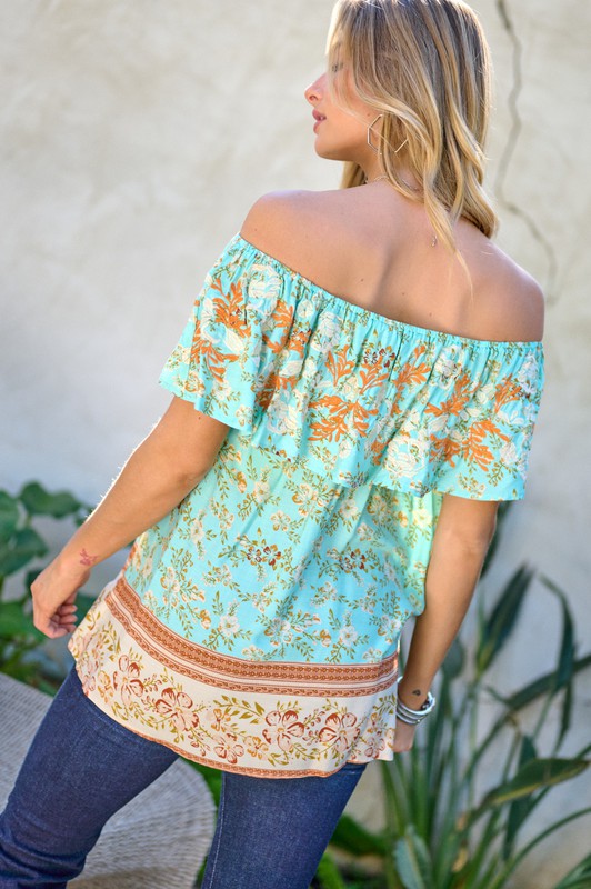 A woman with blonde hair is smiling while standing outdoors, wearing the Printed Off Shoulder Smocked Top paired with jeans.