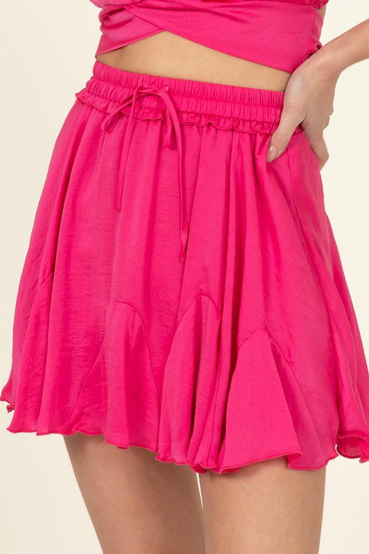 A person is partially visible in the image wearing the Not Your Girl Drawstring Ruffled Mini Skirt, featuring a bright pink flared silhouette with an elastic waistband and drawstring, paired with a matching top.