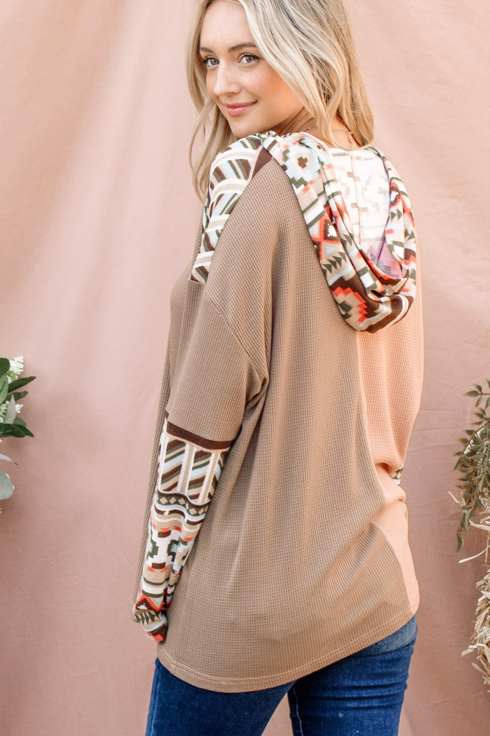 A woman with long blonde hair wears an And The Why Printed Color Block Hooded Waffle Top in various earth tones, standing in front of a light brown background.