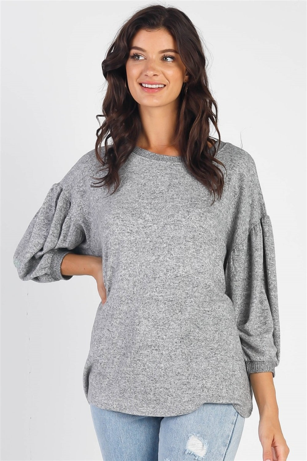 A person with long dark hair stands against a white background, smiling and raising one hand while wearing the trendy and stylish Cherish Apparel Drop Shoulder Puff Sleeve Top in burgundy paired with light blue jeans.