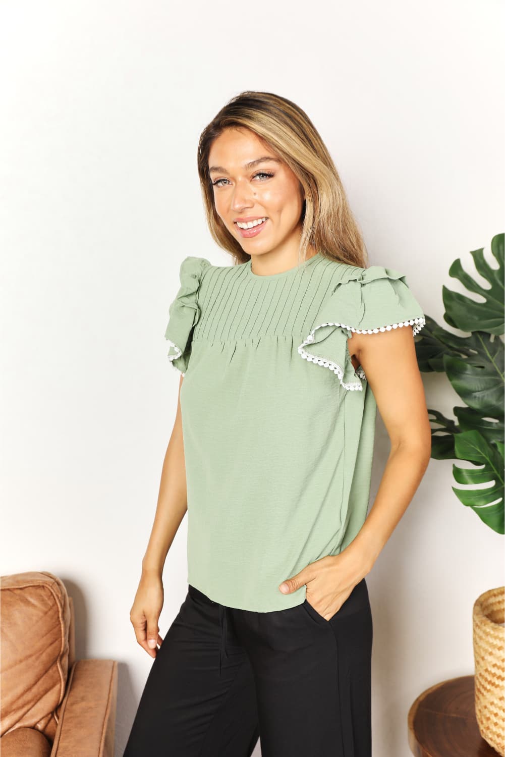 A woman stands and smiles, wearing the Perfee Pleated Detail Flutter Sleeve Blouse in light green paired with black pants.