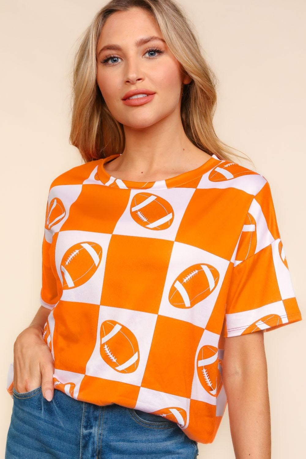 A woman wears a Haptics Football Checkered Print Short Sleeve T-Shirt in orange and white, standing against a plain background, and pairs it perfectly with denim shorts for a casual sporty look.