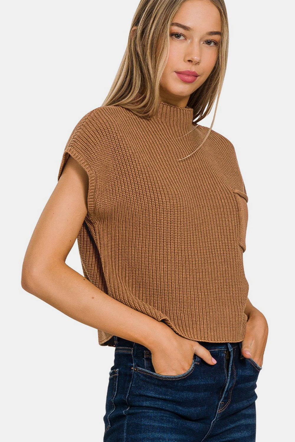 A person with long hair is wearing a Zenana Mock Neck Short Sleeve Cropped Sweater in brown and blue jeans. They are touching their hair with one hand and looking to the side, showcasing the casual comfort of their attire.