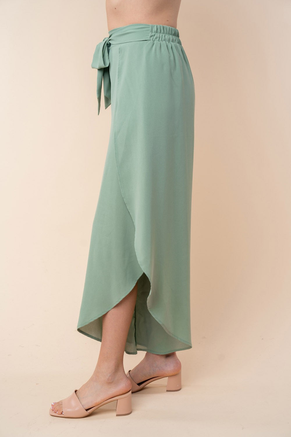 A woman wears White Birch high-waisted woven pants in a flowy green fabric, showcasing a tulip hem and a charming front tie bow, excellently paired with beige open-toe sandals.