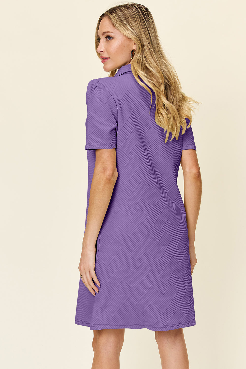 A woman wearing the Double Take Full Size Texture Collared Neck Short Sleeve Dress, featuring a geometric pattern in purple and made from soft polyester, stands against a neutral background. She has long, light hair and is looking slightly to the side. The dress is available in sizes S-3XL and is easy to care for—just machine wash cold.