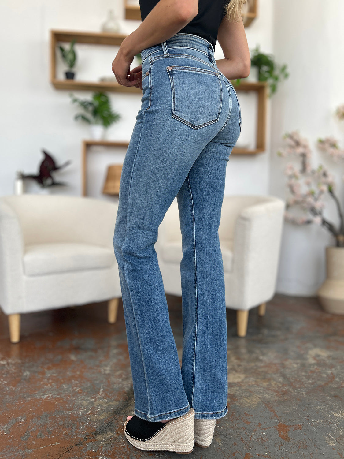 Person wearing Judy Blue Full Size Mid-Rise Waist Straight Jeans and a black top, standing in a room with white chairs, potted plants, and wooden shelves. Only the lower half of the body and part of the arms are visible.