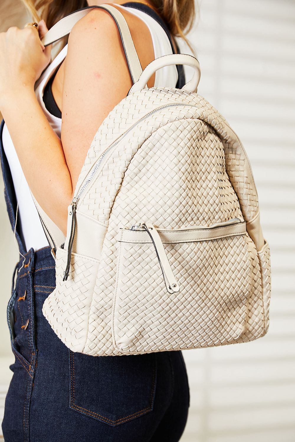 A woman stands indoors smiling, carrying a light-colored SHOMICO PU Leather Backpack on her shoulder while wearing a dark sleeveless top and jeans.