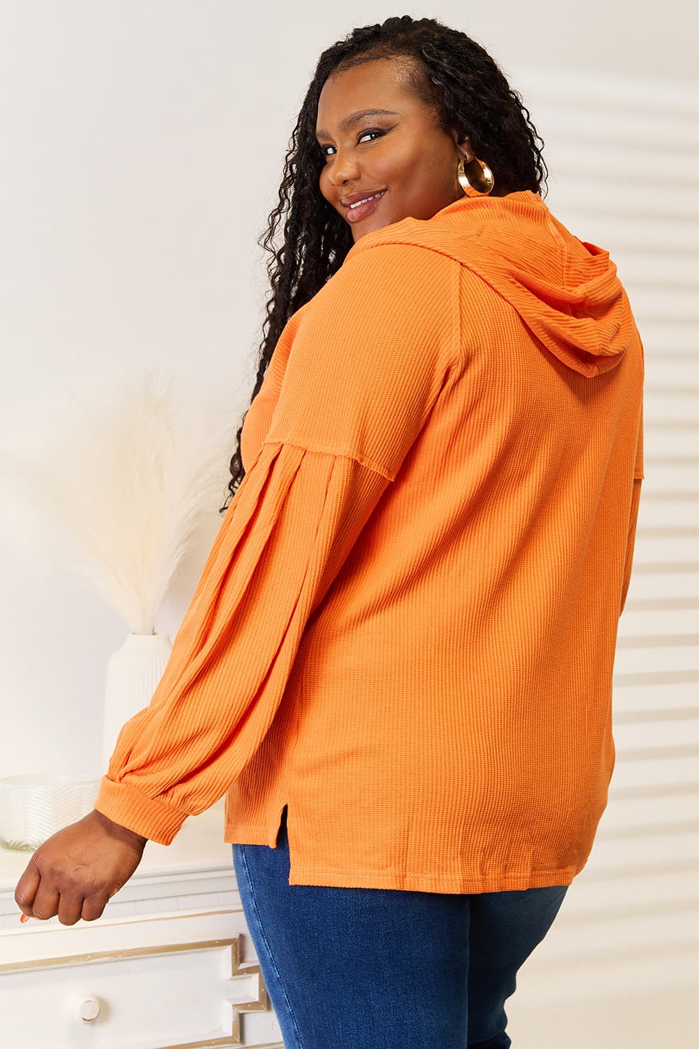 A person wearing an orange Basic Bae Side Slit Buttoned Waffle Knit Hoodie with blue jeans stands indoors near a white dresser, smiling and looking to the side.