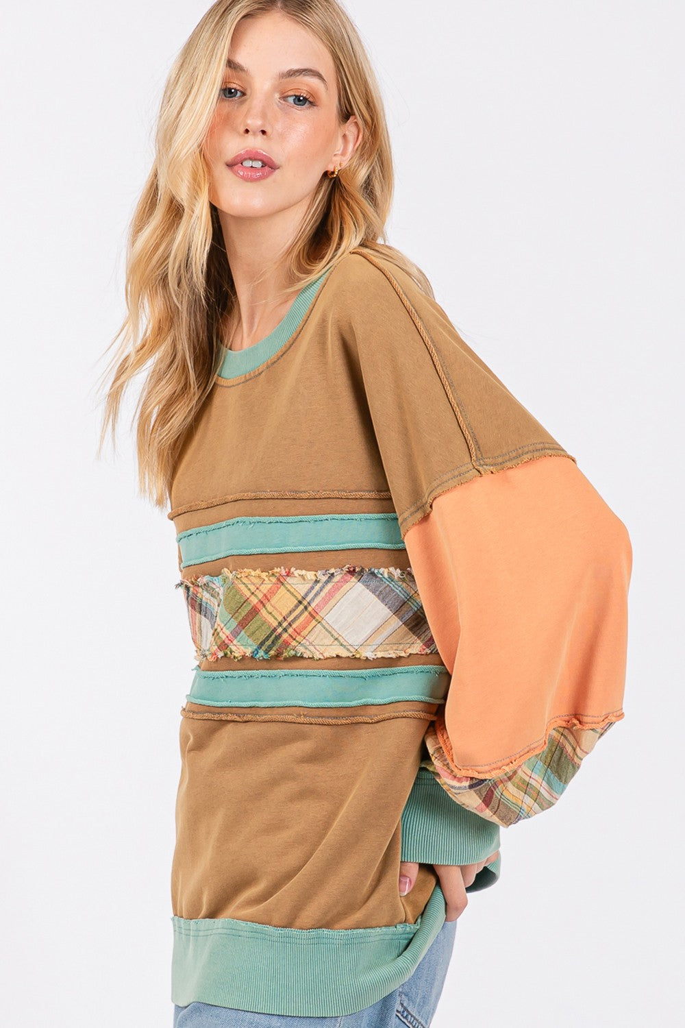 A person wearing a brown hat and the SAGE + FIG Mineral Wash Raw Edge Color Block Sweatshirt, featuring a patchwork design with plaid print contrast and solid color panels, smiles while posing with one hand in their pocket.