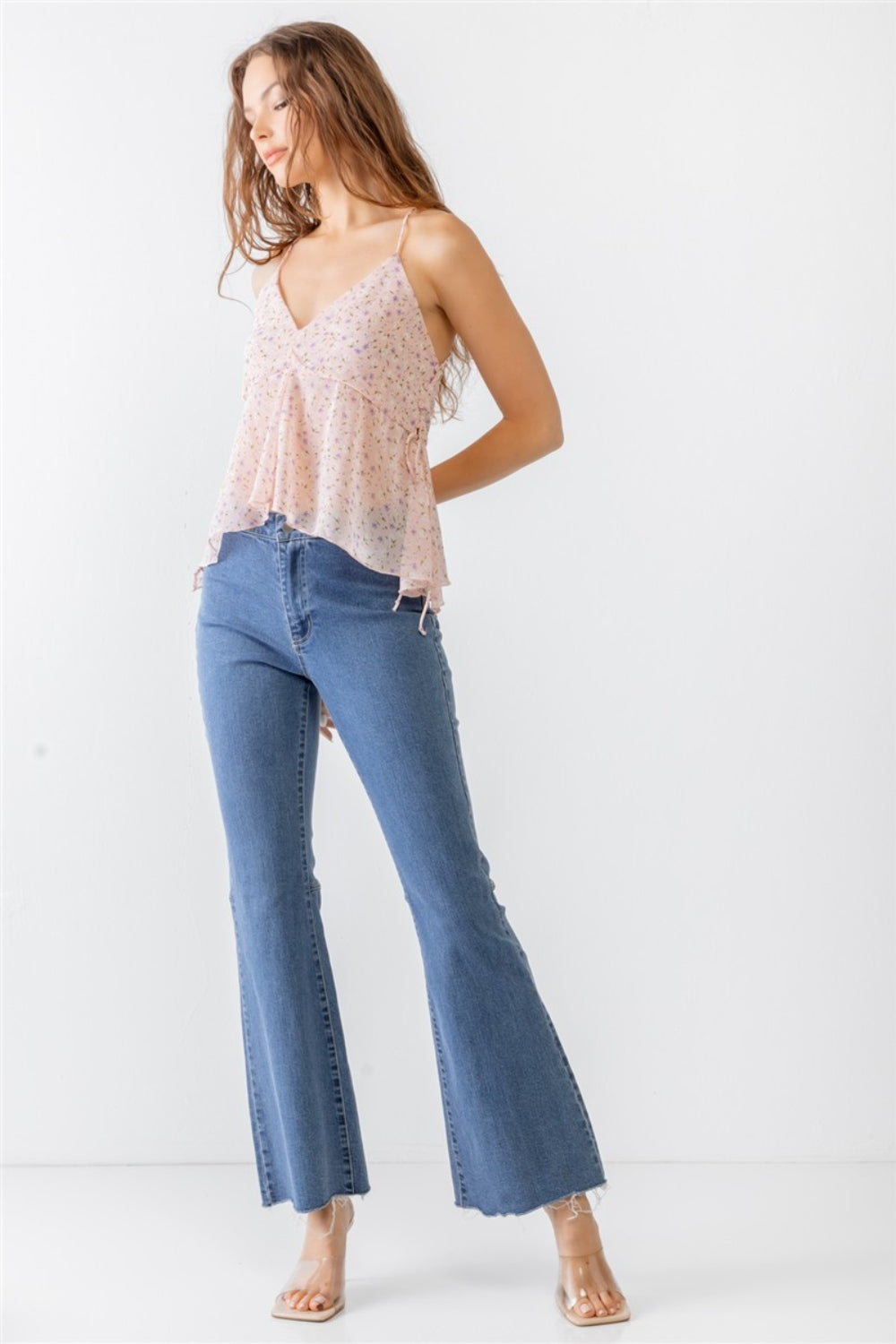 A woman wearing the Miss Love / Kevin Co. Floral Print V-Neck Criss-Cross Back Flare Cami and blue jeans stands against a plain white background.