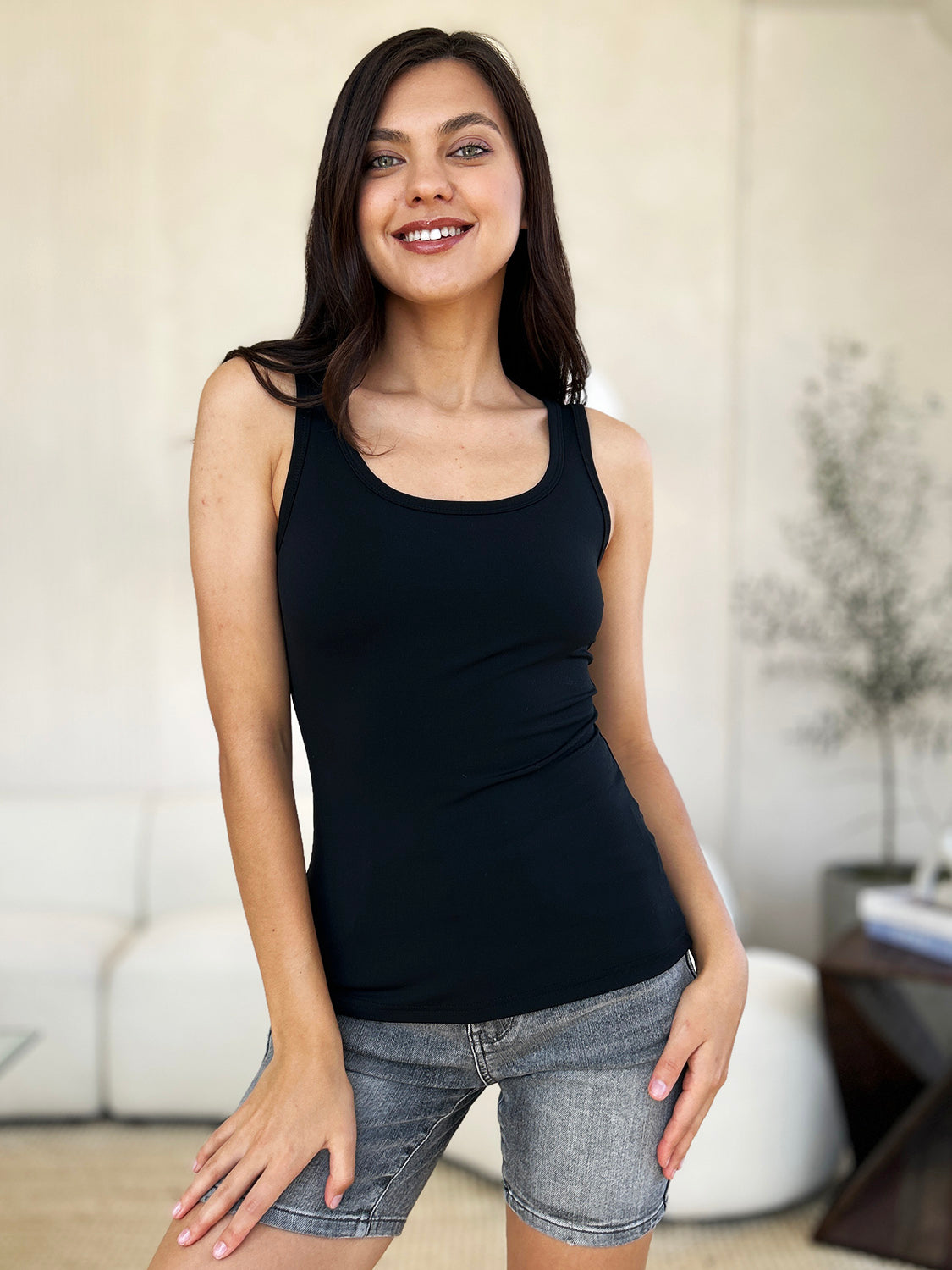 A woman with brown hair, wearing sunglasses, blue jeans, and the Basic Bae Full Size Square Neck Wide Strap Tank in black stands against a concrete wall, showcasing a basic style.