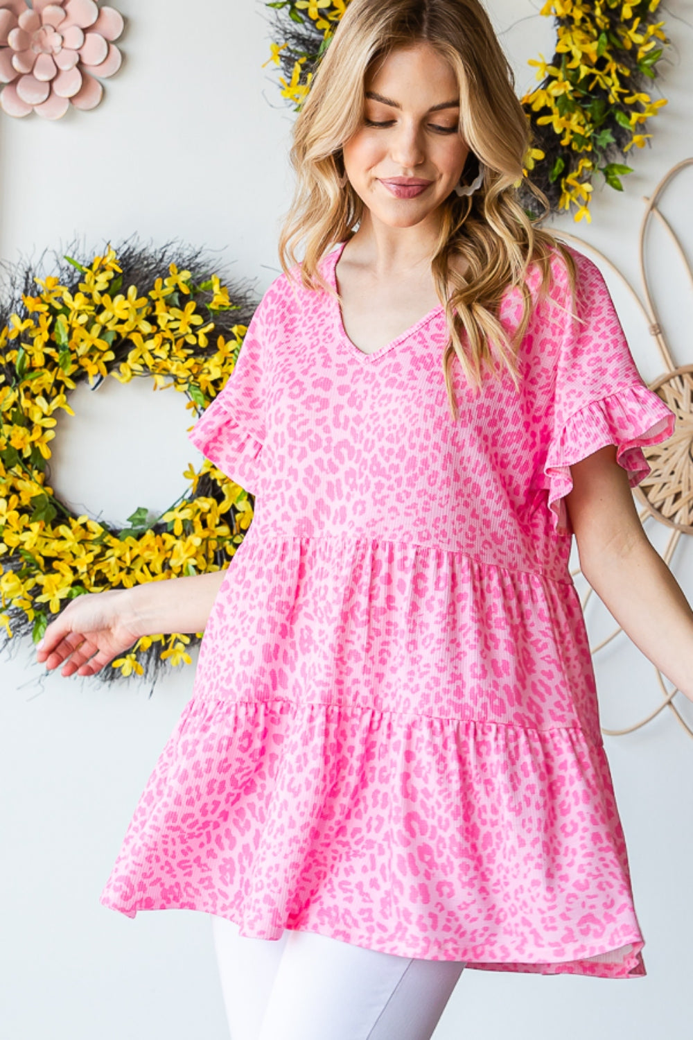 A person wearing a Heimish Full Size Leopard Ruffle Trim Short Sleeve Top stands in front of a floral wreath display.