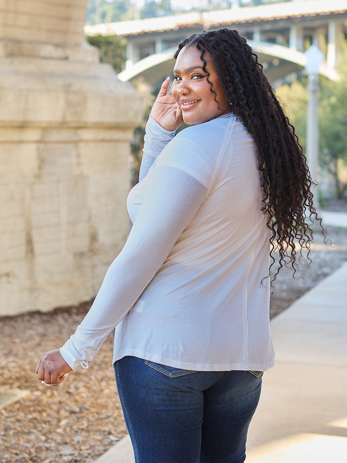 A woman wearing sunglasses and a slightly stretchy Basic Bae Full Size Round Neck Long Sleeve T-Shirt in white stands on a sidewalk with an arched structure in the background, smiling slightly.