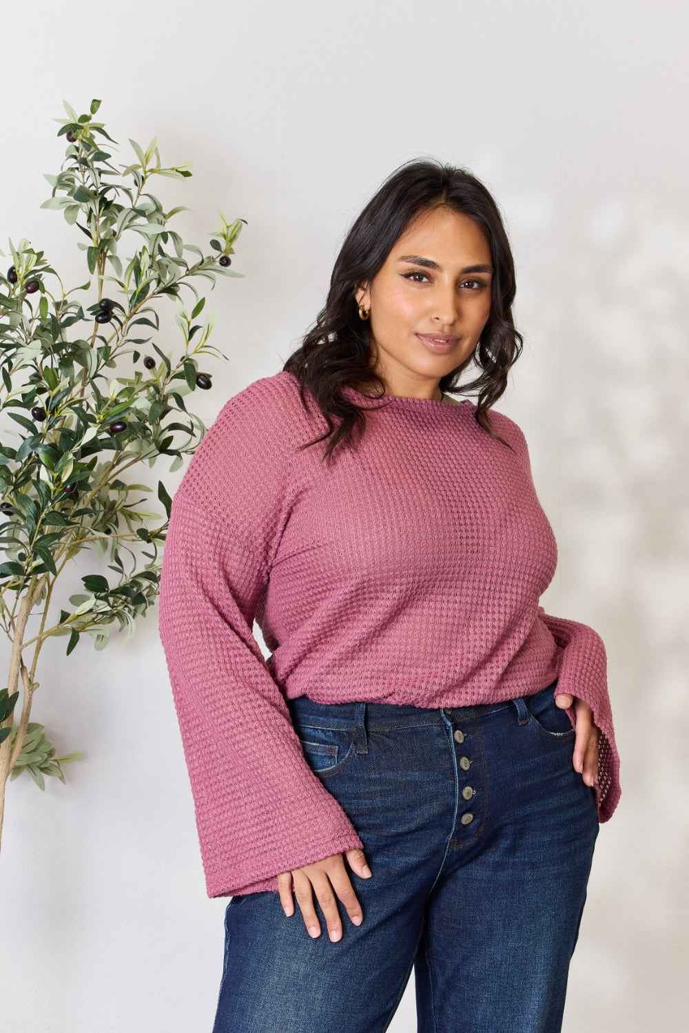 A person wearing a Culture Code Full Size Waffle-Knit Round Neck Long Sleeve Blouse in pink and jeans stands in front of a light background, complemented by a leafy plant on the left.